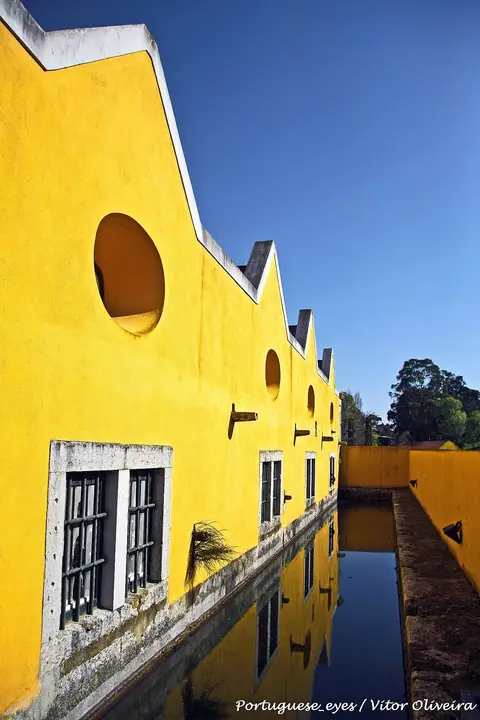 Edifício amarelo da Fábrica da Pólvora de Barcarena refletido num canal de água, sob céu azul.