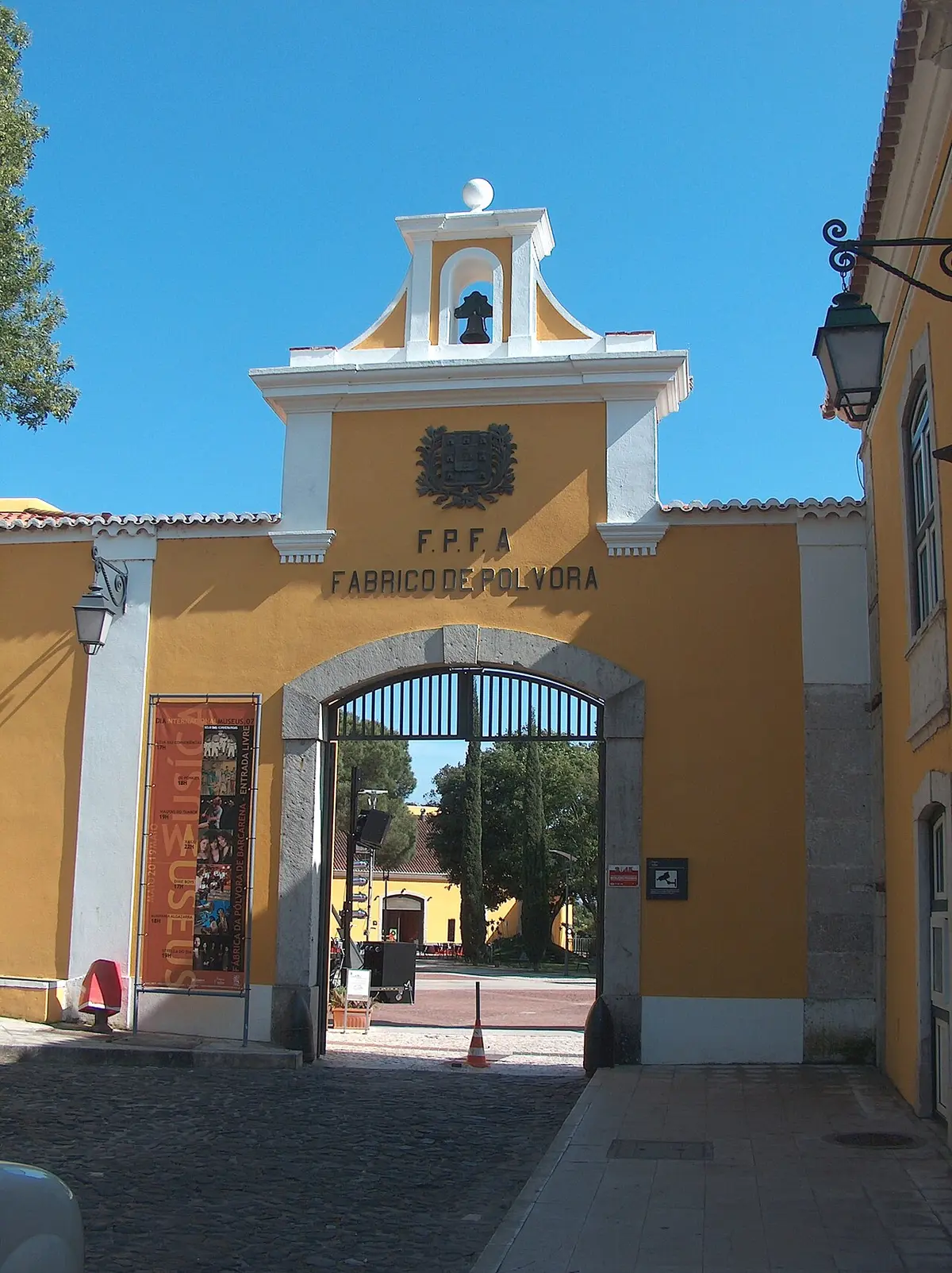 Portão amarelo de entrada da Fábrica da Pólvora de Barcarena, com sineira e brasão.