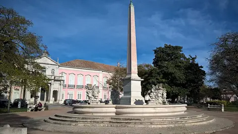 Obelisco e fonte de pedra num jardim, com edifício rosa e árvores ao fundo