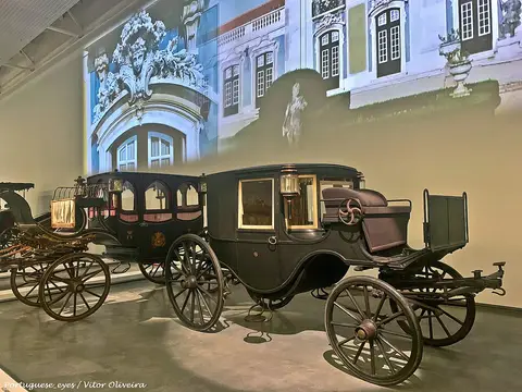 Coches antigos expostos numa sala do Museu Nacional dos Coches, com projeção de fachada palaciana na parede