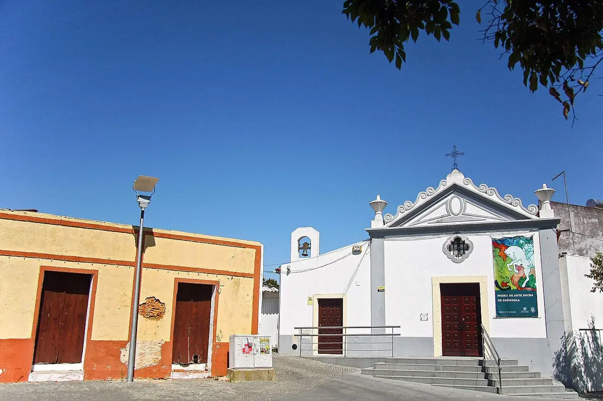 Fachada branca do Museu de Arte Sacra de Grândola, com cruz, campanário e escadaria de acesso