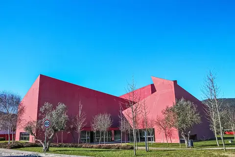Edifício moderno de fachada vermelha da Casa das Artes, rodeado de árvores e relva, sob céu azul