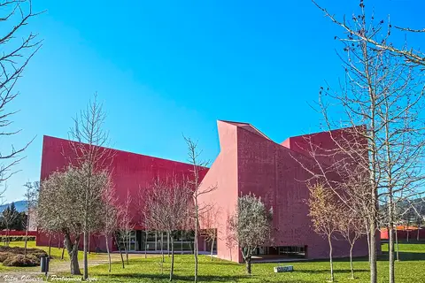 Edifício moderno de fachada vermelha da Casa das Artes, rodeado de relva e árvores, sob céu azul.