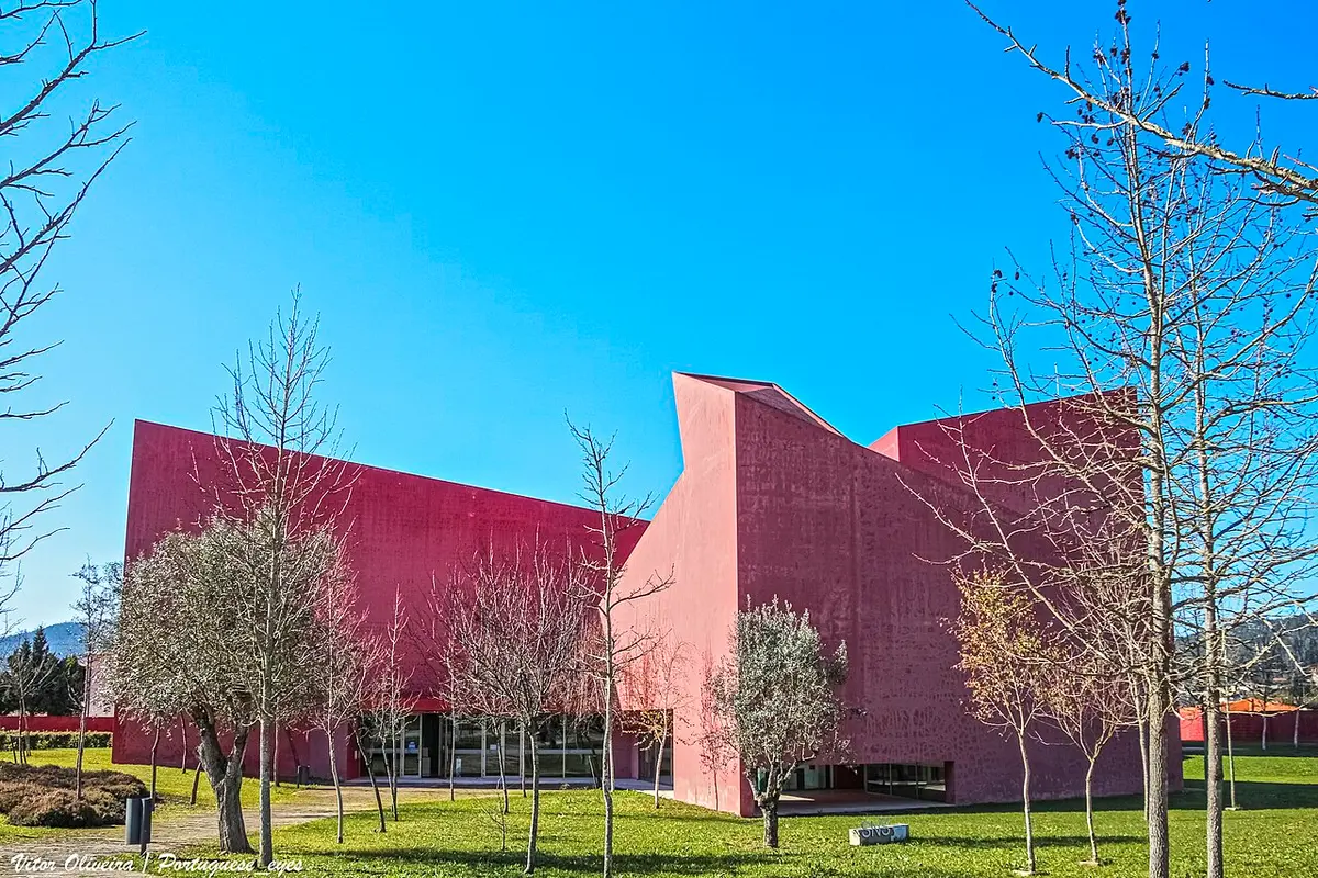 Edifício moderno de fachada vermelha da Casa das Artes, rodeado de relva e árvores, sob céu azul.