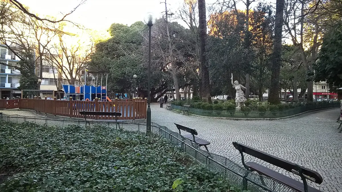 Jardim da Parada com bancos, parque infantil vedado e estátua entre árvores ao fim da tarde
