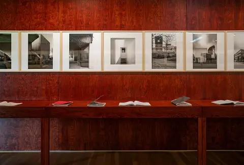 Sala de exposição com paredes de madeira, fotografias emolduradas e livros sobre mesa comprida