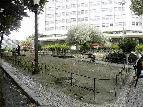Jardim urbano com gradeamento, árvore florida e bancos, junto a edifício moderno