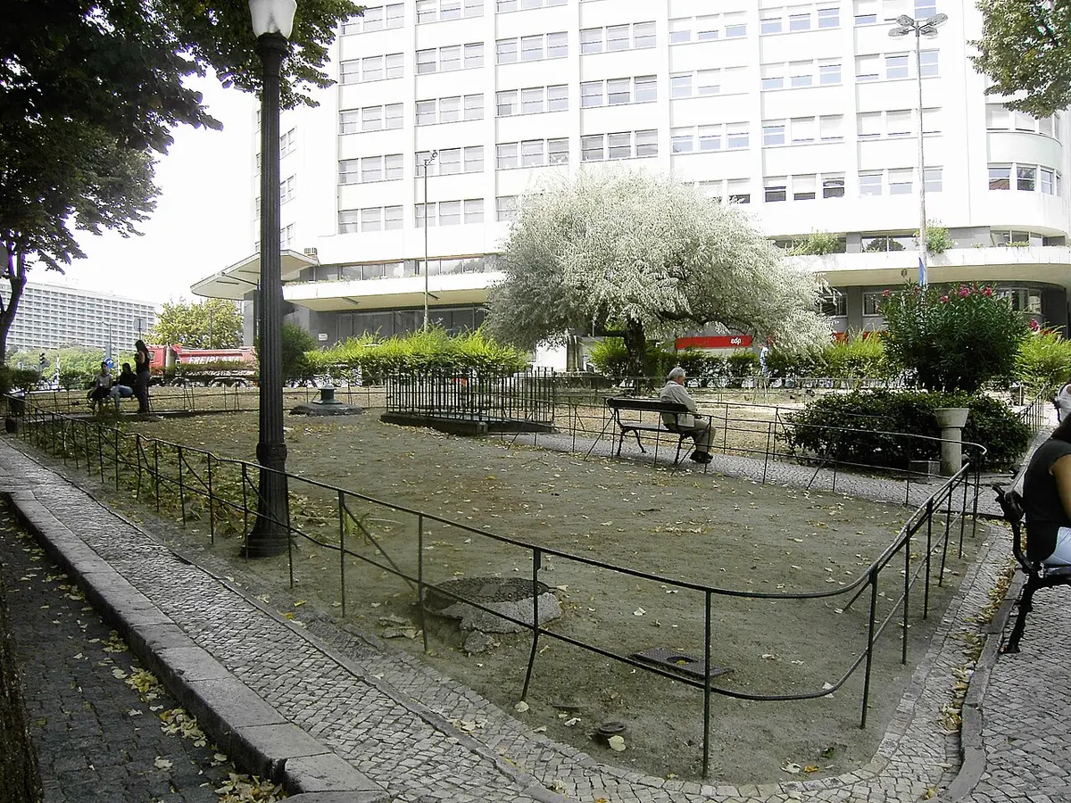 Jardim urbano com gradeamento, árvore florida e bancos, junto a edifício moderno