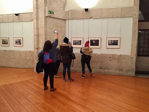 Sala de exposições com fotografias emolduradas nas paredes de pedra e visitantes de costas