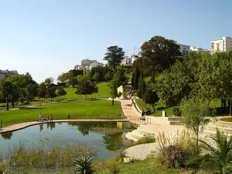 Parque urbano com lago, relvados e árvores, com prédios ao fundo em Lisboa