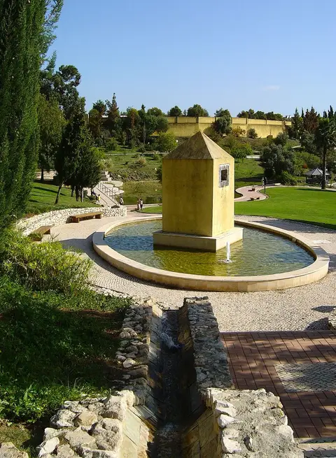 Fonte circular com torre amarela num parque ajardinado com relvados, árvores e caminhos empedrados.