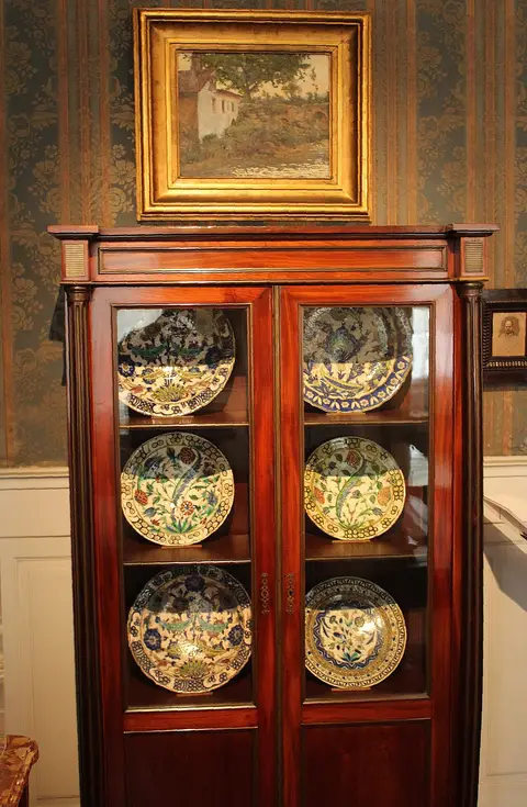 Vitrina de madeira com pratos de faiança decorados e quadro a óleo na parede, em casa-museu.