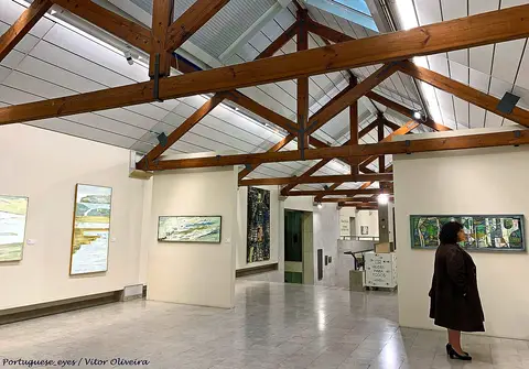 Sala de exposições do museu com telhado de vigas de madeira e quadros nas paredes brancas