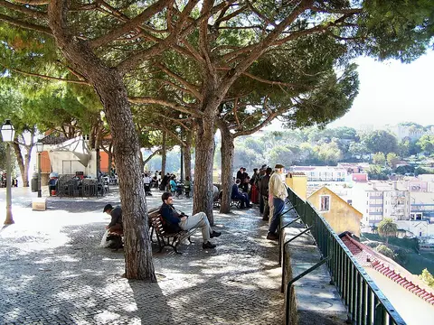 Miradouro da Graça com pinheiros, esplanada e visitantes junto à grade, com vista sobre Lisboa