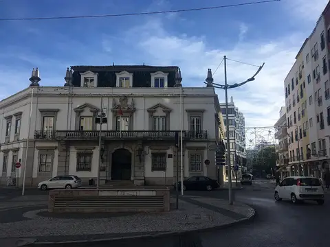 Edifício histórico de dois pisos numa praça de Faro, com carros e passeio calçado em frente.