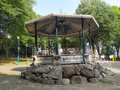 Coreto de ferro sobre base de pedra, rodeado de árvores, num parque urbano.