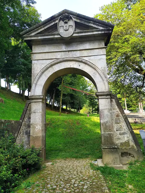 Arco de pedra com brasão à entrada do Parque da Ponte, rodeado de relva e árvores
