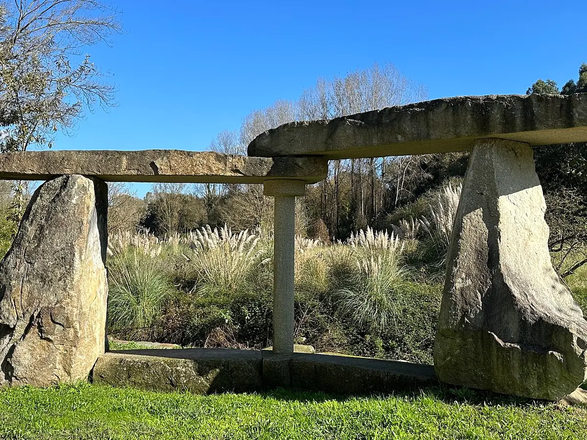 Estrutura de pedra granítica sobre relva num parque, com canas e vegetação ao fundo e céu azul.