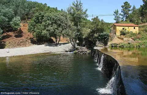 Praia fluvial com açude e pequena queda de água, margem de seixos e casa amarela entre árvores.