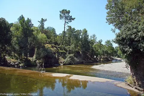 Praia fluvial dos Moinhos de Alva, com rio de águas calmas, margem de seixos e árvores