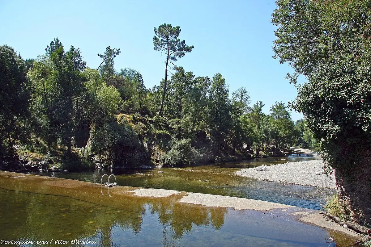 Praia fluvial dos Moinhos de Alva, com rio de águas calmas, margem de seixos e árvores