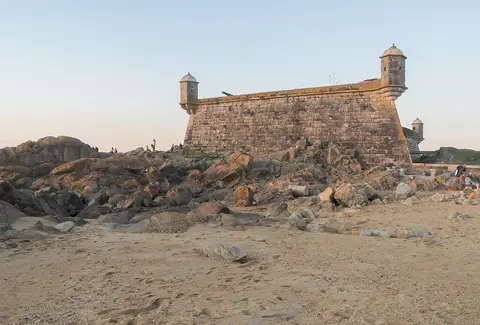 Castelo do Queijo visto da praia, com muralhas de pedra, guaritas e rochedos ao pôr do sol.