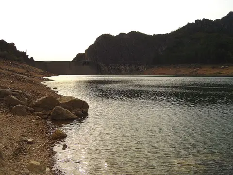 Albufeira de Santa Luzia com margem de pedras, água calma e a barragem ao fundo entre encostas rochosas