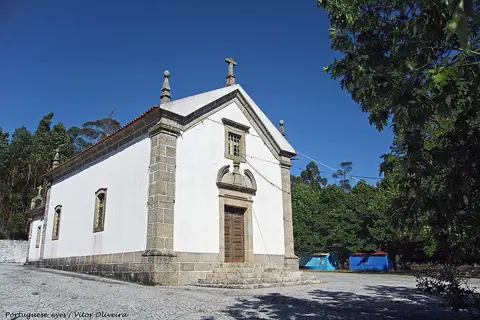 Capela branca da Senhora da Ribeira, com cantaria de granito e cruz, rodeada de árvores sob céu azul