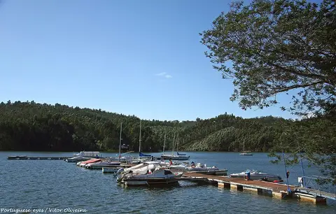 Pontão com pequenas embarcações atracadas numa albufeira rodeada de encostas arborizadas