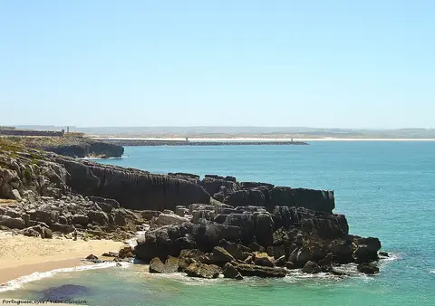 Praia de areia junto a formações rochosas escuras e mar azul-turquesa em Peniche
