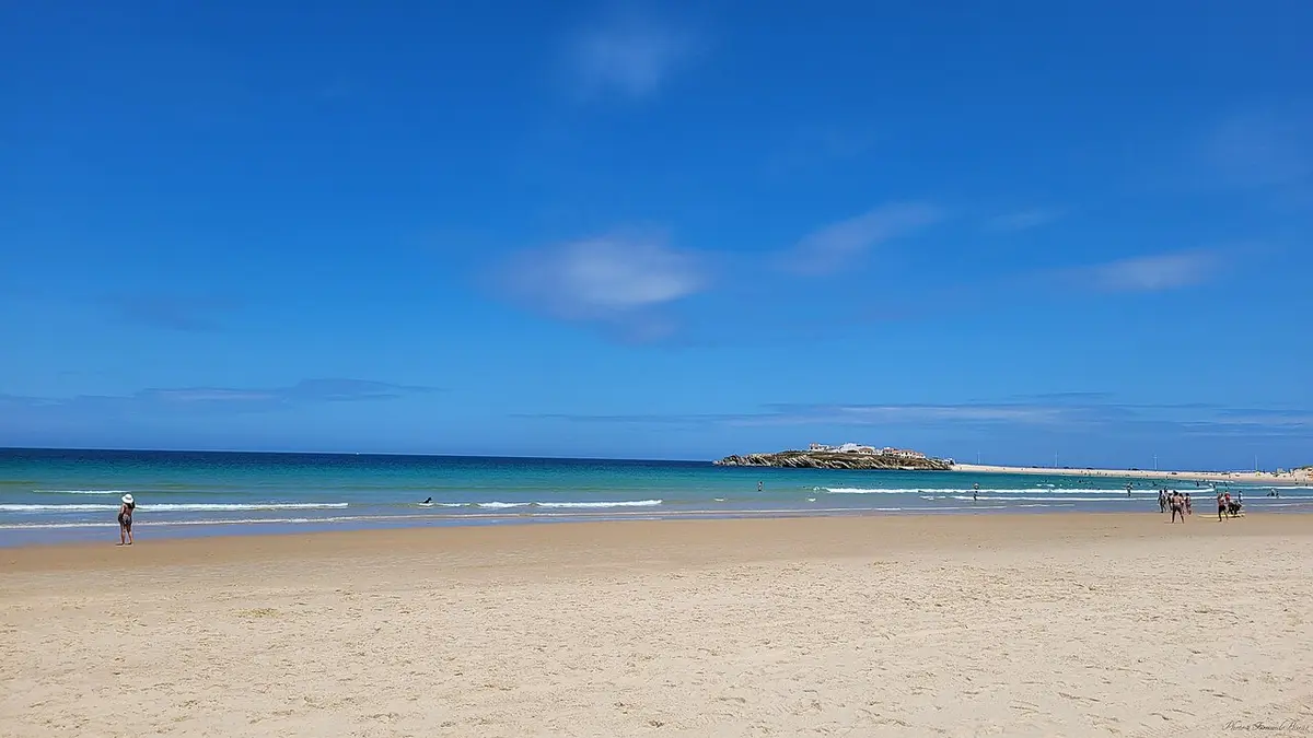 Praia de areia larga com mar azul e a ilha do Baleal ao fundo, sob céu limpo.