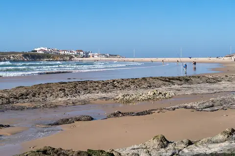 Praia do Baleal com rochas na areia, ondas e casas brancas do Baleal ao fundo, sob céu azul.