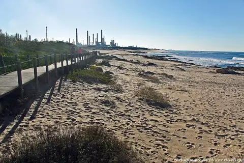 Passadiço de madeira junto à praia de areia do Cabo do Mundo, com refinaria ao fundo