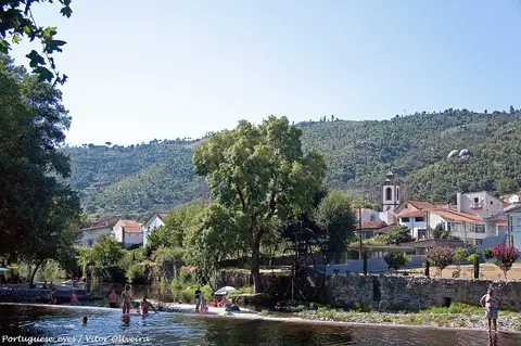 Praia fluvial de Sandomil com rio, árvores, azenha e aldeia com igreja ao fundo