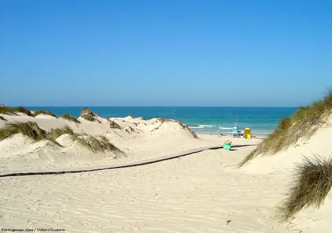 Praia da Cova da Alfarroba com dunas, passadiço de areia e mar azul ao fundo