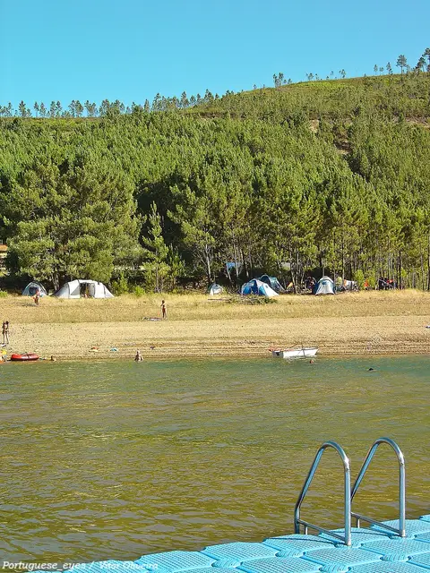 Praia fluvial de Fernandaires na albufeira de Castelo de Bode, com tendas entre pinheiros e pontão azul.