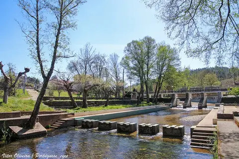 Praia fluvial do Açude do Pinto com passadiço de pedras sobre o rio, árvores e céu azul