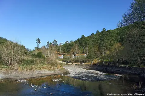 Praia fluvial do Pego das Cancelas, com rio de águas calmas, margem de seixos e encosta arborizada