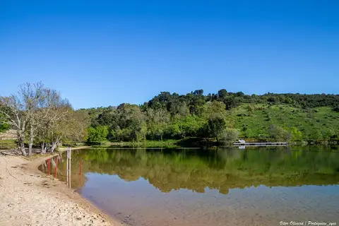 Praia fluvial com areia e água calma rodeada de árvores sob céu azul