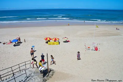 Praia da Pedra do Ouro com areal largo, chapéus de sol coloridos e posto de vigilância junto ao mar.