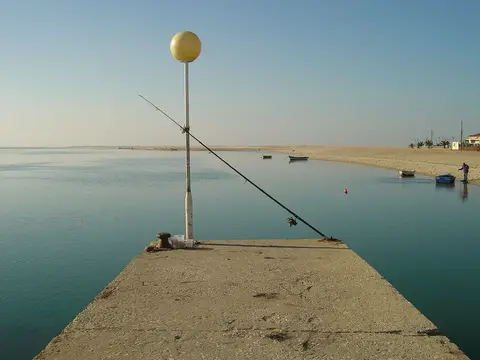 Pontão de betão com cana de pesca sobre água calma da lagoa, com barcos e areal ao fundo