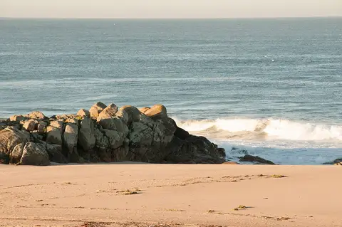 Praia de areia com formação rochosa e ondas a rebentar no mar ao fim da tarde