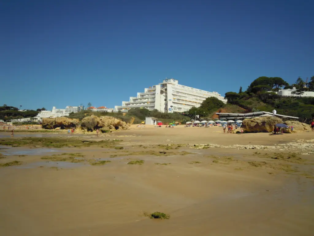 Praia da Oura em Albufeira, com areal largo, rochas e edifícios hoteleiros ao fundo sob céu azul