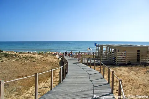 Passadiço de madeira sobre dunas a descer para a praia da Galé-Oeste, com mar e apoio de praia.