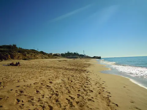 Areal extenso da Praia da Galé com falésias baixas ao fundo e mar calmo sob céu azul.