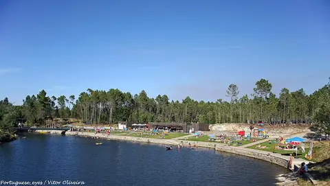 Praia fluvial do Trabulo com relvados, parque infantil e banhistas junto à albufeira rodeada de pinhal.