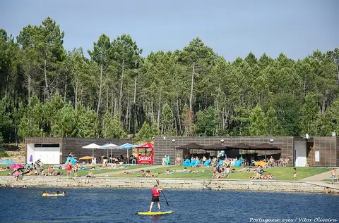 Praia fluvial do Trabulo com relvado, bar de apoio e pinhal; banhistas e prática de paddle no rio.