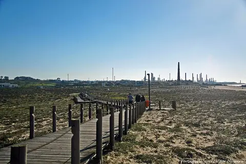 Passadiço de madeira sobre dunas junto à praia, com o horizonte industrial ao fundo