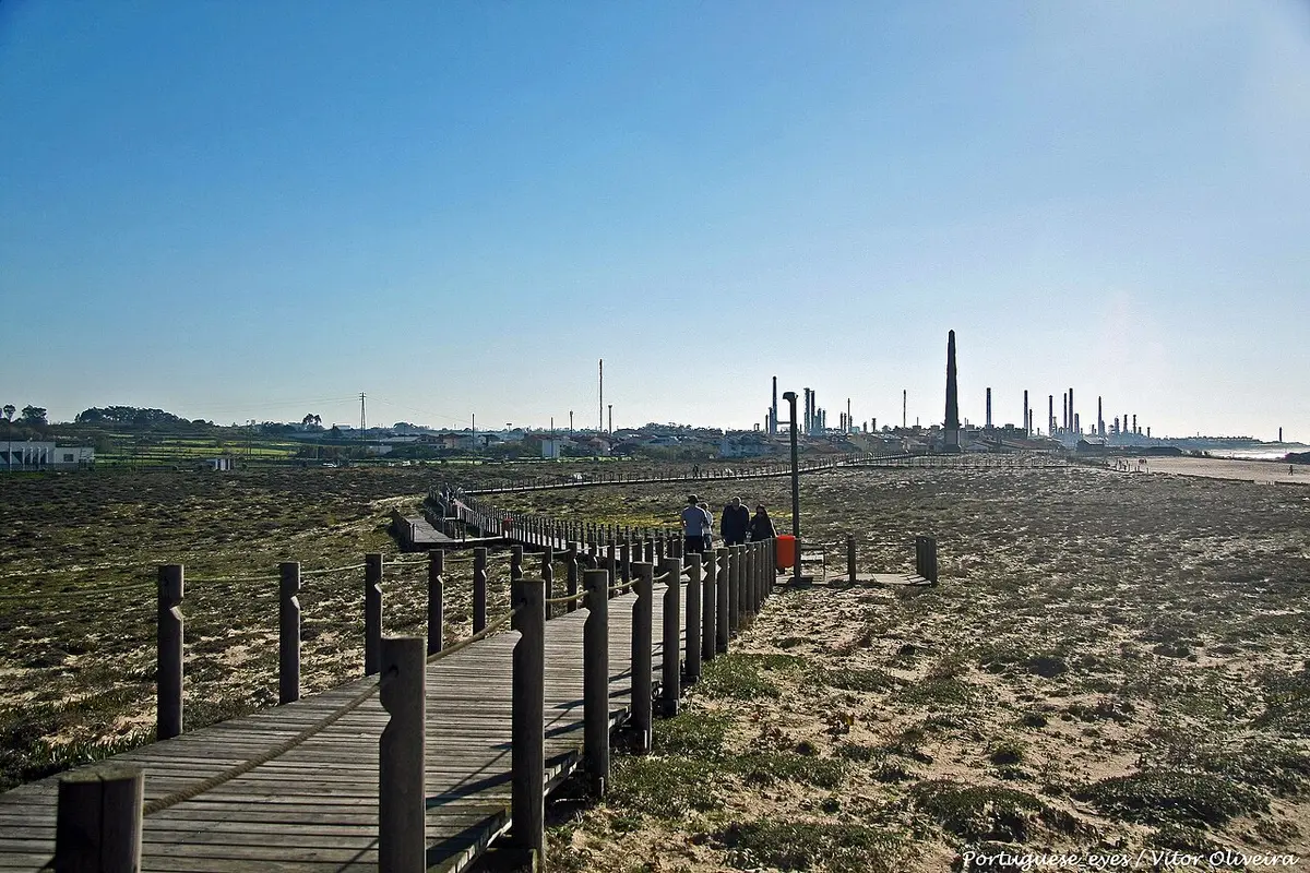Passadiço de madeira sobre dunas junto à praia, com o horizonte industrial ao fundo