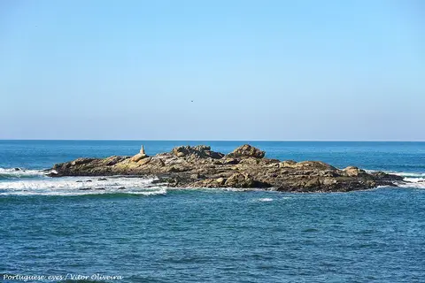 Ilhéu rochoso ao largo da Praia do Marreco, rodeado por mar azul e ondas, sob céu limpo